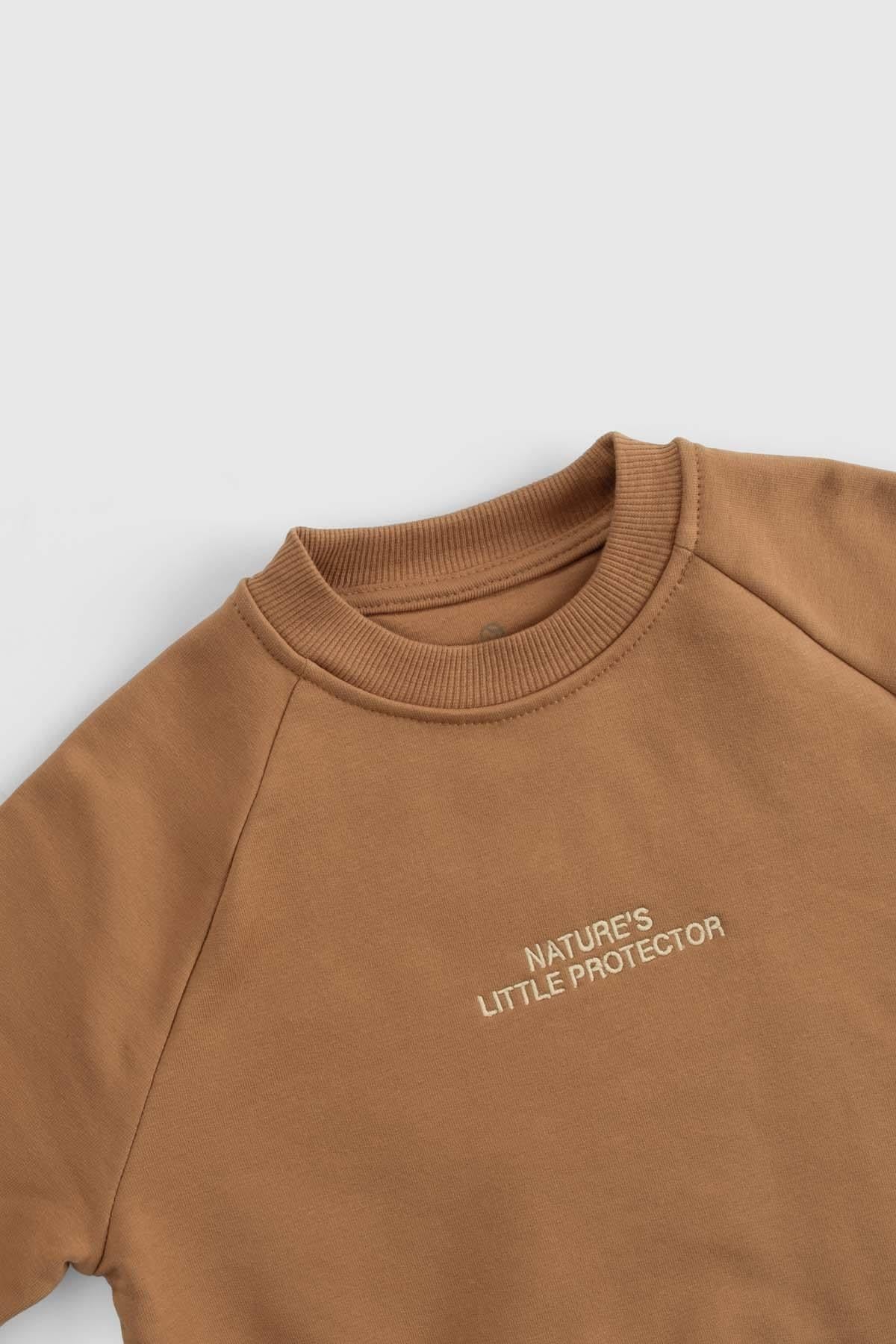 Close-up of organic cotton baby tracksuit top featuring 'Nature's Little Protector' embroidery in brown.